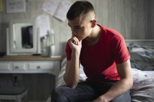 Pilot mental health programme for young people: Teenage boy sitting in his bedroom looks anxious as he rests his head in his hand
